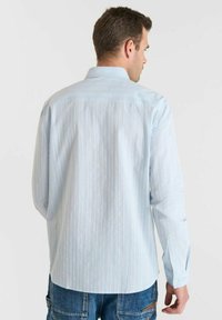 Man in een lichtblauw gestreept overhemd met lange mouwen en blauwe jeans, gezien van achteren tegen een effen witte achtergrond.