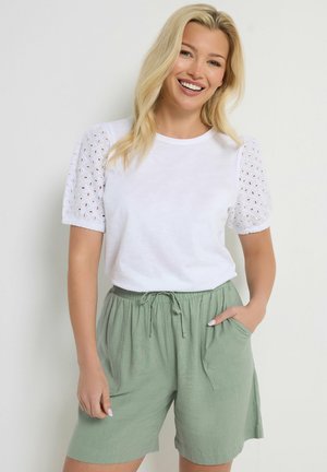 Donna sorridente con lunghi capelli biondi che indossa una maglietta bianca a maniche corte con occhielli e pantaloncini verdi chiaro con coulisse, in piedi con una mano in tasca.