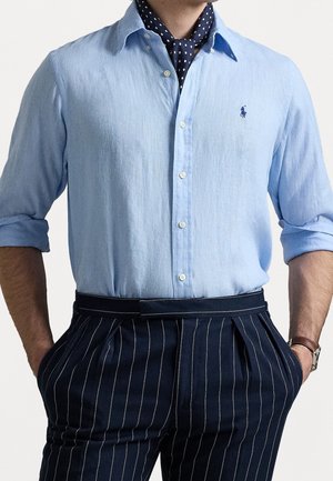Homme portant une chemise bleu clair à boutons avec manches retroussées, une écharpe bleu marine à pois, un pantalon bleu marine foncé à fines rayures, une montre et un bracelet en cuir.