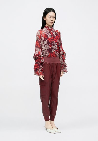 Áttetsző piros virágos blúz fodros ujjakkal, bordó cargo nadrággal, melyen zsebek és elasztikus mandzsetták találhatók, valamint fehér hegyes orrú balerinacipőkkel.