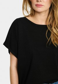 Sort kortærmet t-shirt lavet af ribbet stof, med bred hals og afslappet pasform. Blød struktur med opkrøllede ærmeafslutninger.