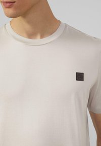 Lichtbeige t-shirt van zachte stof, met een ronde halslijn en een klein donker vierkant logo op de linkerborst.