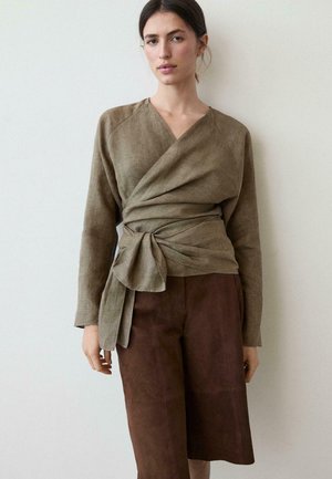 Kobieta w długim, oliwkowym topie typu wrap, związanym w talii, i brązowych spodniach do kolan, stojąca na tle gładkiej jasnej ściany.