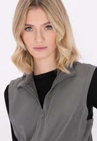 Gilet in pile grigio con colletto alto e cerniera frontale completa, caratterizzato da una consistenza morbida e senza motivi visibili. Indossato sopra un top nero.