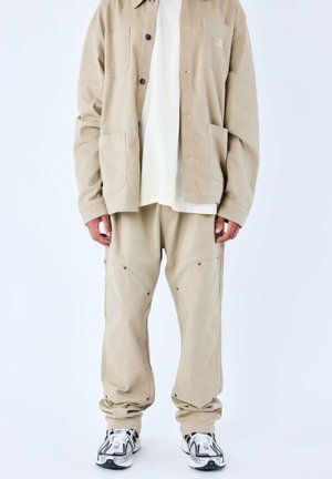 Veste en toile beige avec deux poches avant sur une chemise blanche, associée à un pantalon beige ample avec des poches à angle, porté avec des baskets.