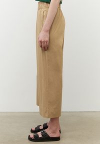 Beige, löst sittande beskurna byxor med elastisk midja och sidofickor, i kombination med svarta sandaler med dubbla spännen.