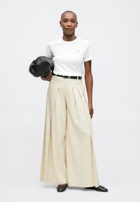 Žena v bílém tričku a béžových širokých plisovaných kalhotách, drží černou tašku, stojí před jednoduchým světlým pozadím.