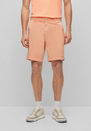 Mann trägt hellorange Shorts, passendes oranges Hemd, weiße Socken und beige-weiße Sneakers, steht vor einem einfarbigen grauen Hintergrund.