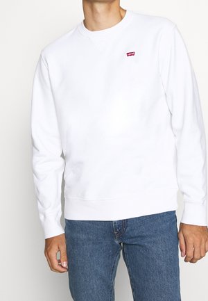 Homme portant un sweat-shirt Levi’s blanc à col rond et un jean bleu, debout les bras détendus le long du corps devant un fond uni.