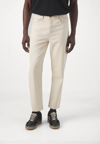 Jeans beige claros con un diseño de pierna recta, que presentan bolsillos frontales y cierre de cremallera. Combinados con zapatillas negras y una camiseta oscura.