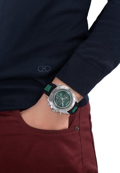 Kronografur med klockhus i rostfritt stål, grön urtavla, svart gummirem och silverdetaljer. Bärs på en handled med en marinblå tröja.