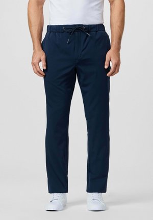 Hombre vestido con pantalones azul marino con cordón y zapatillas blancas, de pie frente a un fondo gris liso.