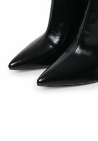 Zapatos de tacón alto de charol negro con punta afilada y un acabado brillante, que presentan una textura suave y líneas limpias.