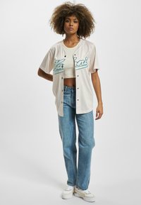 Lys beige knappet skjorte med teal bokstaver og svarte aksenter, båret over en crop top, kombinert med falmede blå jeans og hvite sneakers.