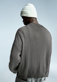 Grå ribbet striktrøje med løs pasform og faldende skuldre, ledsaget af en hvid ribbet beanie. Baggrunden er en blød blå gradient.
