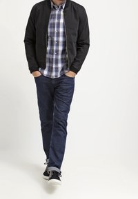 Mustat pommikoneetakki sinivalkoisen ruutupaidan päällä, yhdistettynä tummiin farkkuihin ja mustiin lenkkareihin, joissa on valkoiset pohjat.