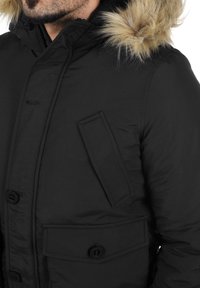 Sort parka med hætte trimmet med pels, knaplukning og en brystlomme. Har en glat tekstur og et holdbart materiale.