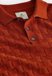 Chemise polo couleur rouille avec un motif carré texturé, trois boutons sombres et une étiquette de marque portant le nom "NEXT" sur le col.