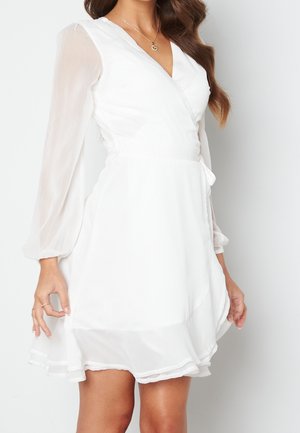 Femme portant une robe portefeuille blanche avec des manches longues transparentes et un ourlet à volants, accessoirisée d'un pendentif et de bagues.
