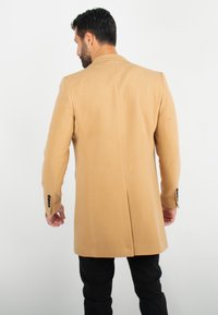 Homme aux cheveux et à la barbe noirs portant un manteau long couleur chameau et un pantalon noir, debout de dos sur fond blanc.