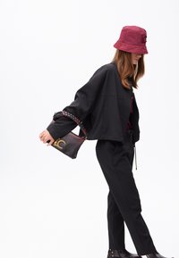 Una donna con una giacca nera corta e pantaloni aderenti indossa un cappello a secchiello bordeaux, tenendo in mano una borsa con una tracolla decorativa.