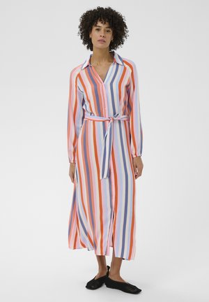 Ženska, oblečena v obleko z dolgimi rokavi in pasom z navpičnimi oranžnimi, roza, modrimi in belimi črtami ter črnimi natikači, stoji na belem ozadju.