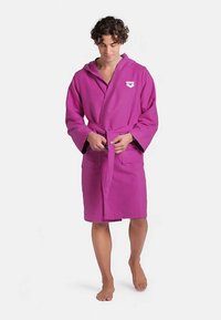 Accappatoio in cotone viola con cappuccio, maniche lunghe, cintura in vita e tasche frontali. Presenta un logo bianco sul petto. Tessuto morbido e dalla trama strutturata.