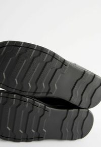Semelles de chaussures noires avec une adhérence à motif, présentant des rainures profondes et des surfaces texturées pour une meilleure traction et stabilité.