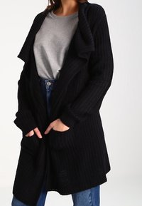 Cardigan noir en tricot avec une coupe ample, un grand col et des poches à l'avant. Porté sur une chemise grise et un jean bleu.