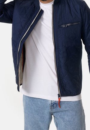 Mann trägt eine geöffnete marineblaue gesteppte Jacke über einem weißen T-Shirt und hellblauen Jeans vor weißem Hintergrund.