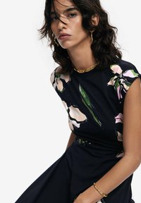 Schwarzes Kleid mit kurzen Ärmeln, ausgestattet mit einem floralen Druck in Pink und Grün. Glattes Material, figurbetonte Silhouette, akzentuiert mit goldener Schmuck.