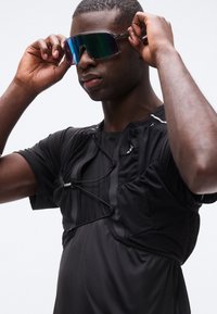 Αθλητικό άτομο που ρυθμίζει μεγάλα αντανακλαστικά αθλητικά γυαλιά ηλίου, φορώντας μαύρο διχτυωτό και υφασμάτινο αθλητικό γιλέκο πάνω από μαύρο πουκάμισο.