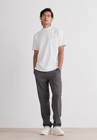 T-shirt bianco a maniche corte con colletto alto, abbinato a pantaloni grigio scuro e sneakers bianche, su uno sfondo chiaro e semplice.