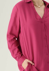 Camicia fucsia con colletto e maniche lunghe, caratterizzata da una texture liscia, bottoni visibili e tasche laterali.