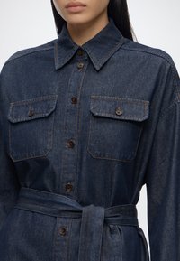 Femme portant une robe-chemise en denim foncé avec des boutons marron, des poches poitrine à rabats, un col pointu et une ceinture en tissu assortie nouée à la taille.
