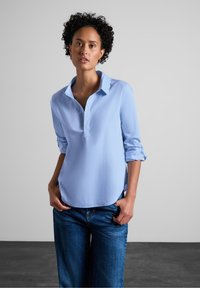 Camicia azzurro chiaro, con bottoni e una texture morbida, maniche lunghe arrotolate. Presenta un colletto e un orlo curvo, abbinata a jeans blu.