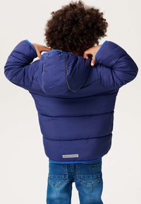 Granatowa kurtka puchowa z kapturem, posiadająca pikowany wzór, niebieską podszewkę oraz metkę "NPPS OUTDOORS" na dolnej części. Noszona z dżinsami.