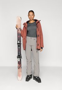 Suusaülikond, mis koosneb roostekarva jope, halli pika varrukaga pluusi, helehallide pükste ja mustade suusasaabastega, hoides roosa ja tumeda suusaga, millel on kinnitused.