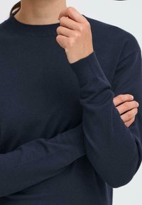 Granatowy sweter dzianinowy z okrągłym dekoltem, ściągaczami na rękawach i gładką fakturą, z założonymi rękami i luźnym krojem.