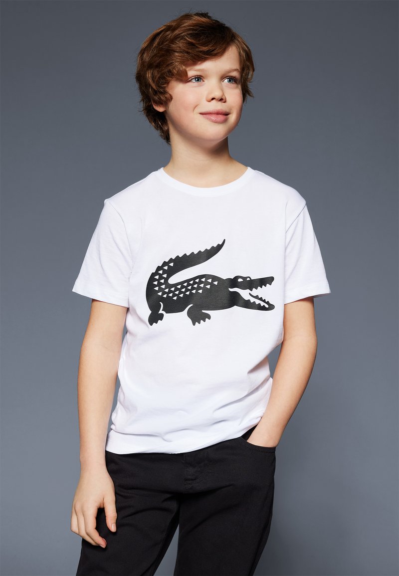 T-shirt en coton blanc avec un grand motif de crocodile noir aux motifs géométriques. Le modèle porte un pantalon noir, les mains dans les poches.
