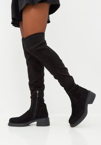 Botas negras de ante por encima de la rodilla con cremallera lateral y tacón de goma negro grueso. Textura suave y diseño ajustado.