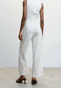 Persona vestida con un mono blanco sin mangas con finas rayas verticales grises y sandalias de tacón marrón oscuro, de pie sobre un suelo claro.