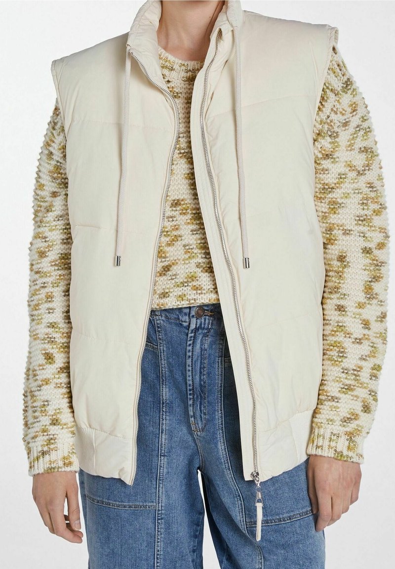 Lehká béžová bezrukávová bunda s vysokým límcem, vybavená předním zipem a šňůrkami na stažení. Nositá přes texturovaný, barevně kombinovaný svetr.