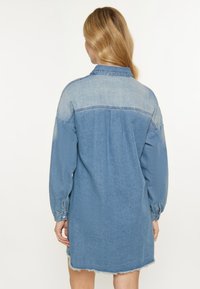 Denim-paitamekko vaaleansinisen ja tummansinisen sävyissä. Varustettu kauluksella, pitkillä hihoilla ja repaleisella helman elämyksellä. Pehmeä tekstuuri, rento istuvuus.