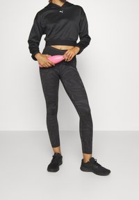 Felpa nera corta con un piccolo logo bianco, abbinata a leggings scuri e decorati. Il modello tiene in mano un attrezzo per il fitness rosa e indossa scarpe da ginnastica nere.