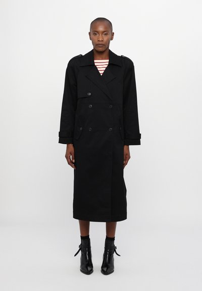 Abrigo negro de trench con diseño de doble botonadura, solapas anchas y bolsillos laterales. Lleva puesto sobre una camiseta a rayas, combinado con botas negras hasta el tobillo.