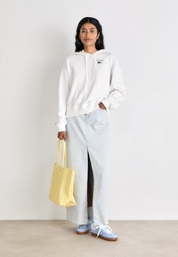 Hanorac alb cu glugă și logo verde, fustă din denim albastru deschis cu slit frontal, geantă de tote galbenă, adidași alb-albastri cu talpă maro.