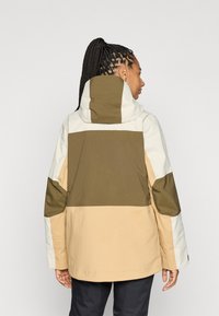Värgblocekterad jacka med paneler i beige, olivgrön och kräm; texturerad tyg med huva och justerbara funktioner, utformad för funktionalitet.