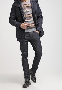 Parka navy con cappuccio staccabile, maglione lavorato a maglia a motivi in grigio, marrone e arancione, jeans blu scuro e scarpe in pelle nere.