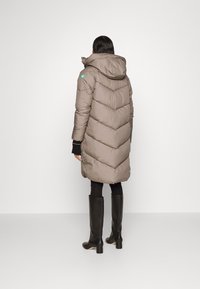 Kvinde iført en lang taupe pufferjakke med hætte, sorte leggings og knæhøje sorte støvler, stående mod en ensfarvet baggrund med ryggen til.